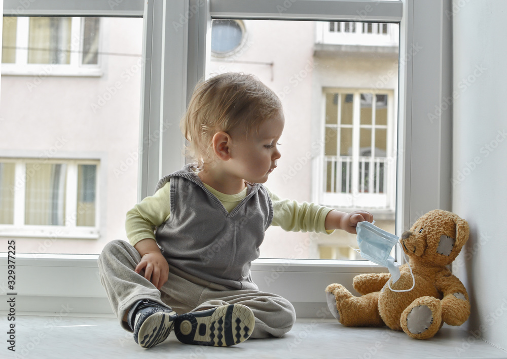 Child in home quarantine at the window putting a medical mask on his sick teddy bear, for protection against viruses during coronavirus COVID-19 and flu outbreak. 